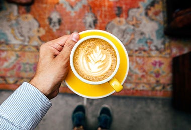 Capuccino con un decorado de flores en su superficie con una persona sosteniendo el plato y la taza de la bebida caliente con leche