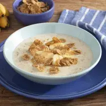 Fotografía en tonos de azul sobre un banco de madera con un paño azul con rayas blancas, un gran plato azul en el centro, con un plato hondo celeste y la papilla con cereales. Al fondo, un racimo de plátanos y una pequeña olla de cereal.