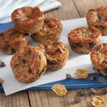 Fotografía en tonos de azul en un banco de madera con un tablero azul, con una hoja de papel encerado y varios muffins de plátano con chispas de chocolate encima. Cereales para el desayuno Nesfit y algunas chispas de chocolate esparcidas por el mostrador.