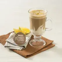 fotografía en tonos blanco y marrón tomada de frente, contiene un paño marrón con un vaso transparente con la bebida de NESCAFÉ Dolce Gusto Café Au Lait con Crema de Leche NESTLÉ. Al lado hay una cápsula de Dolce Gusto y al fondo rodajas de naranja