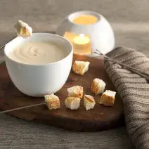 fotografía en tonos grises, marrones y banco visto de frente. Contiene una tabla de madera redonda con un recipiente blanco redondo que contiene la fondue, 4 tenedores de fondue y trozos de pan alrededor.