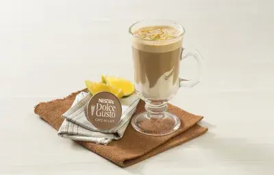 fotografía en tonos blanco y marrón tomada de frente, contiene un paño marrón con un vaso transparente con la bebida de NESCAFÉ Dolce Gusto Café Au Lait con Crema de Leche NESTLÉ. Al lado hay una cápsula de Dolce Gusto y al fondo rodajas de naranja