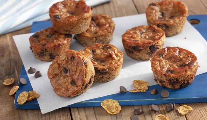 Fotografía en tonos de azul en un banco de madera con un tablero azul, con una hoja de papel encerado y varios muffins de plátano con chispas de chocolate encima. Cereales para el desayuno Nesfit y algunas chispas de chocolate esparcidas por el mostrador.