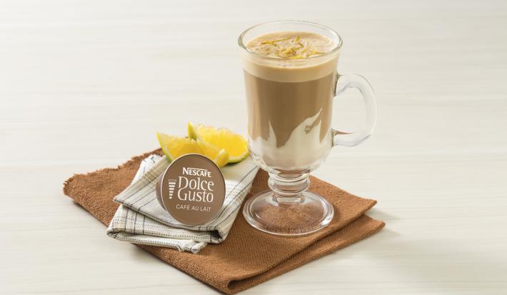 fotografía en tonos blanco y marrón tomada de frente, contiene un paño marrón con un vaso transparente con la bebida de NESCAFÉ Dolce Gusto Café Au Lait con Crema de Leche NESTLÉ. Al lado hay una cápsula de Dolce Gusto y al fondo rodajas de naranja